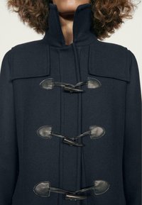 Cappotto di lana blu navy con colletto alto, dotato di chiusure a scorrimento e dettagli in pelle. Tessuto testurizzato con motivo a zigzag.