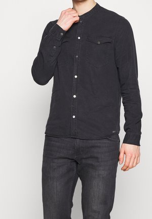 Mand iført en sort skjorte med knapper og brystlommer samt mørke jeans, der justerer kraven med den ene hånd.