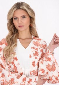 Kevyt kimono, jossa on oransseja kukkakuosia valkoisella taustalla. Syvä V-pääntie valkoinen paita alla. Sileä tekstuuri, leveät hihat.