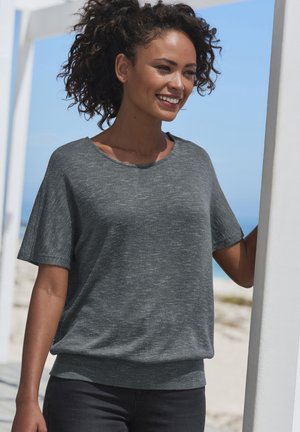 Graues, kurzärmliges Oberteil aus leichtem Stoff, mit lockerer Passform und rundem Ausschnitt. Getragen mit dunklen Jeans vor einem Strandhintergrund.