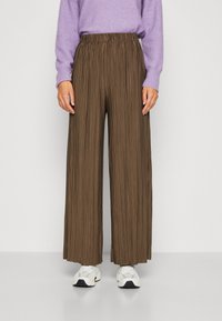Donkerbruine plooibroek met een elastische tailleband, gecombineerd met witte sneakers. De stof heeft een textuur met een verticaal streeppatroon.