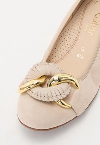 Beige mockask balletskor med rund tå, en vävd rem med en guldfärgad hårdvaruöppning, och en perforerad innersula för andningsförmåga.