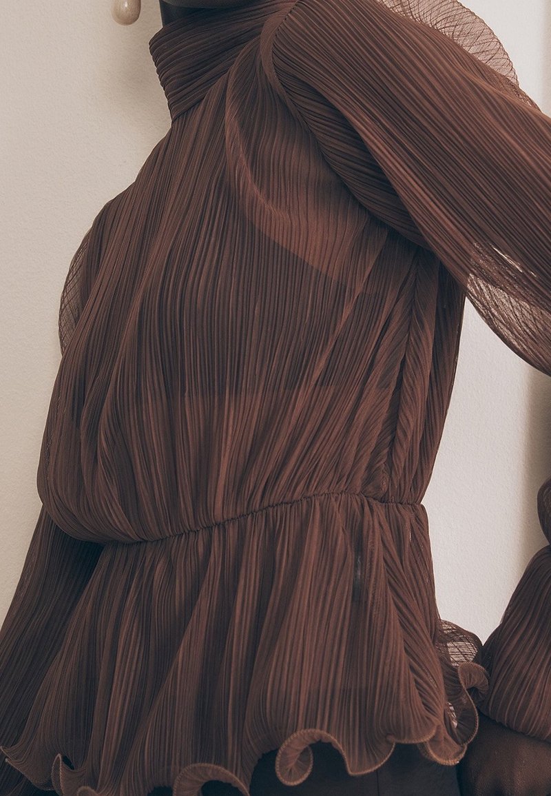 Blouse marron transparente plissée à manches longues, ourlet volanté et col haut, portée par une personne sur un fond clair.