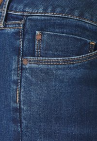 Mörkblå denimjeans med en framficka, som har kontrasterande gyllene sömmar och små metallnitar. Slät textur med en lätt glans.