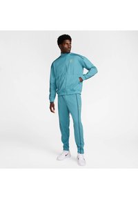 Teal treningsdrakt laget av glatt stoff, med en zip-up jakke og matchende bukser med grå detaljer og en logo på jakken.