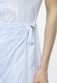 Κοντινή λήψη φωτεινού γαλάζιου και λευκού ριγωτού φορέματος με χρυσή αγκράφα κοντά στη μέση και ορατό γυμνό χέρι ατόμου.