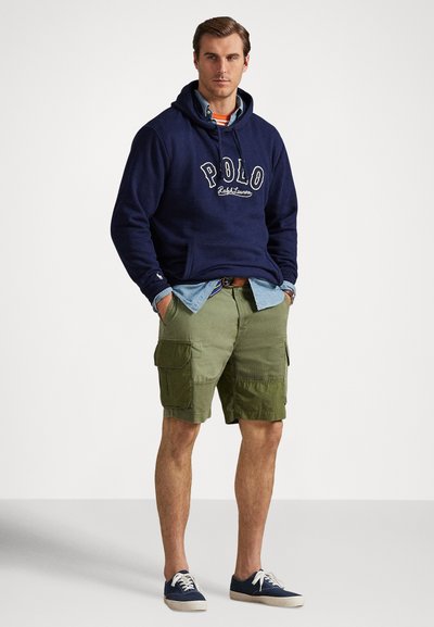 Vyras dėvi tamsiai mėlyną Polo džemperį su gobtuvu, šviesiai mėlyną marškinių palaidinę, oranžinės spalvos dryžuotą marškinėlį po ja, alyvinių tonų kargo šortus ir tamsiai mėlynas sportbačius, stovintis su rankomis kišenėse.