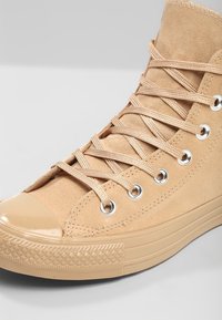 Beige vyšší teniska vyrobená z semiše, s lesklým gumovým špičkovým krytem, kovovými očky a texturovanou gumovou podrážkou s diamondovým vzorem.