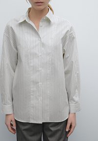 Donna che indossa una camicia bianca con bottoni a righe verticali grigie sottili e pantaloni grigi, in piedi su uno sfondo grigio chiaro uniforme.