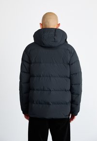 Svart puffjacka med huva, med en quiltad design, framträdande sömmar och elastiska ärmslut. Bakifrån syns materialets textur.