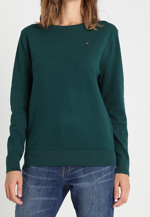 Tummanvihreä, pitkähihaisen pehmeästä materiaalista valmistettu neule, jossa on pyöreä pääntie ja vasemmassa rinnassa logotarra. Yhdistetty sinisiin farkkuihin.