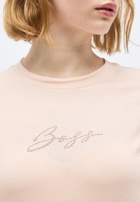 Κοντινό πλάνο ενός ατόμου που φοράει ανοιχτό ροζ πουκάμισο με τη λέξη "Boss" γραμμένη με μικρές στρας στο στήθος.