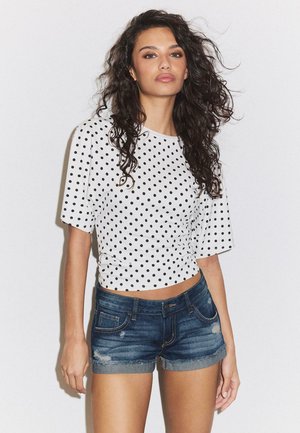 Kvinde med langt, krøllet hår iført en hvid top med korte ærmer og sorte polkaprikker samt slidte denimshorts, der står foran en ensfarvet baggrund.