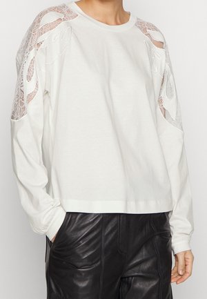 Haut blanc à manches longues avec des découpes en dentelle sur les épaules, associé à un pantalon en cuir noir, les mains dans les poches, debout devant un fond uni.