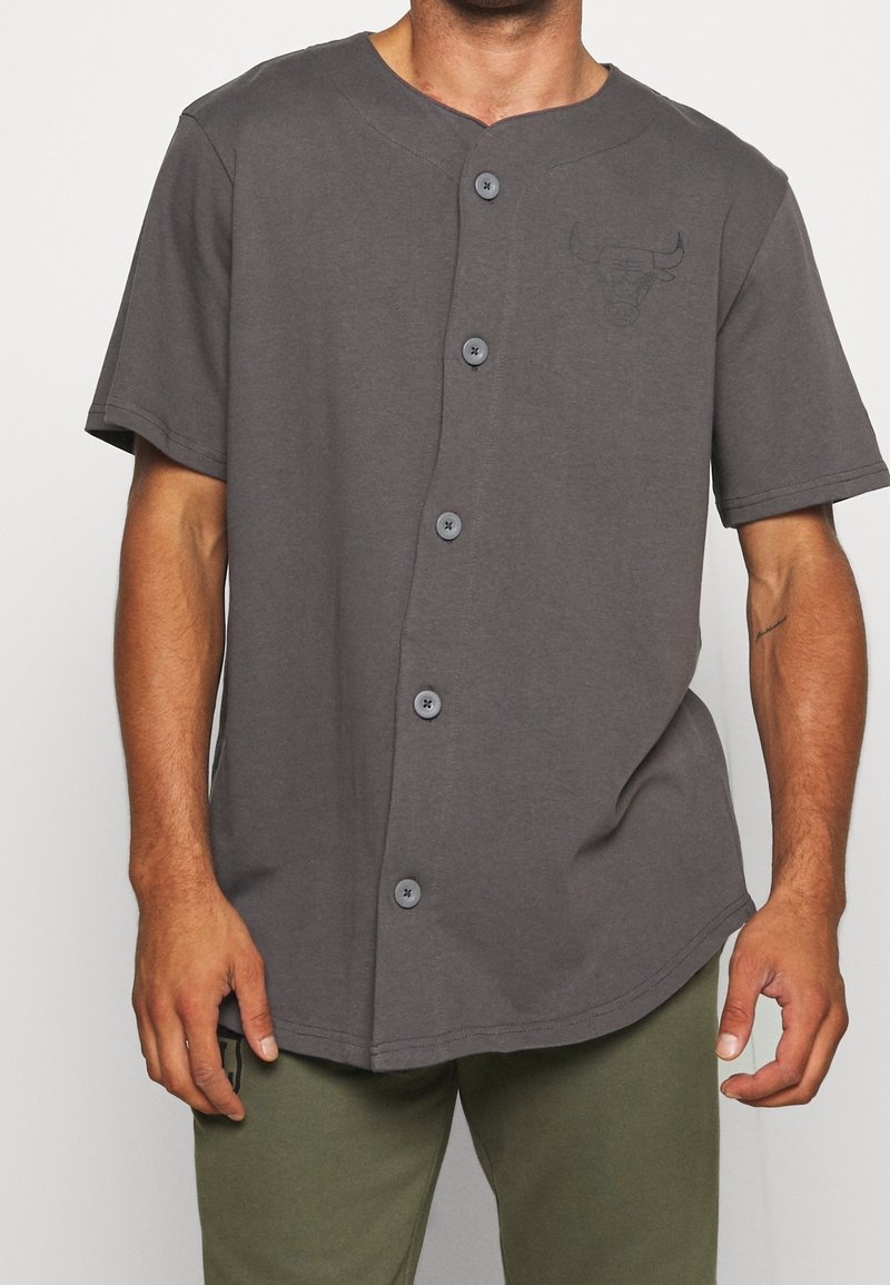 Šedá košile s krátkými rukávy a knoflíky, zaobleným lemem, pěti knoflíky, kulatým výstřihem a nenápadným logem na hrudi.