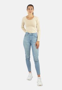 Beige Langarmoberteil und hellblaue hochgeschnittene Jeans, kombiniert mit weißen Sneakern. Das Outfit zeichnet sich durch eine taillierte Passform und eine glatte Textur aus.