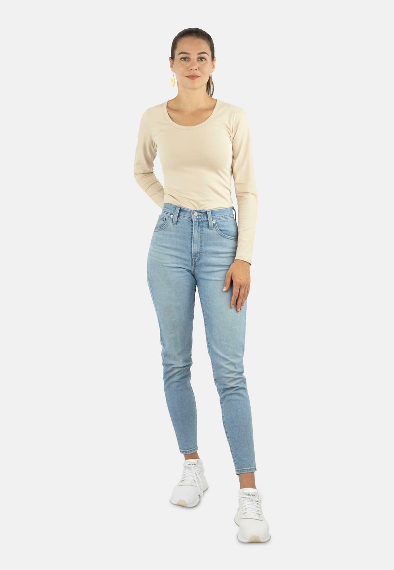 Beige Langarmoberteil und hellblaue hochgeschnittene Jeans, kombiniert mit weißen Sneakern. Das Outfit zeichnet sich durch eine taillierte Passform und eine glatte Textur aus.