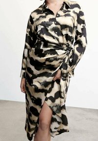 Femme portant une robe boutonnée à manches longues avec un imprimé animal abstrait noir et beige et un détail noué sur le côté, révélant partiellement une jambe.