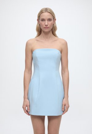 Kobieta o długich blond włosach, ubrana w błękitną mini sukienkę bez ramiączek, stojąca twarzą do przodu na jednolitym białym tle.