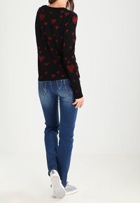 Zwarte trui met rood hartpatroon, lange mouwen, gecombineerd met blauwe jeans en grijze sneakers, met een achteraanzicht van de outfit.