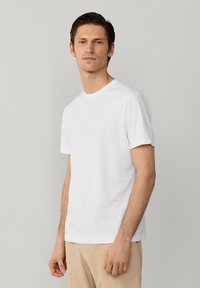 Mann mit dunklen Haaren trägt ein einfaches weißes kurzärmliges Hemd und beige Hosen, steht vor einem hellgrauen Hintergrund und blickt nach vorne.