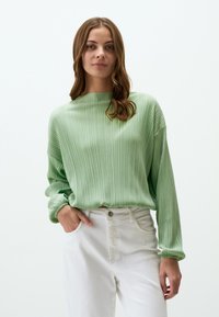 Maglione verde a coste con scollatura ampia e maniche lunghe, abbinato a pantaloni bianchi a vita alta. Texture liscia e vestibilità rilassata.