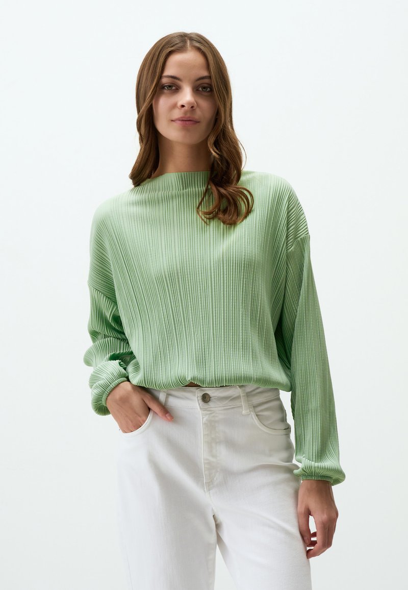 Maglione verde a coste con scollatura ampia e maniche lunghe, abbinato a pantaloni bianchi a vita alta. Texture liscia e vestibilità rilassata.