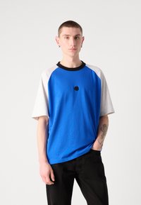 Blue short-sleeve shirt with gray raglan sleeves, black collar, and a black circular accent at the center. Smooth fabric texture.