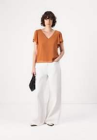 Blusa de cuello en v color óxido con mangas de vuelo, combinada con pantalones anchos blancos. La modelo sostiene un pequeño bolso negro. Acabado de tela suave.