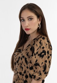 Hnědá blůza s černým leopardím potiskem, se zdobenými rukávy a výstřihem do V. Modelka nosí velké perlové kroužkové náušnice.