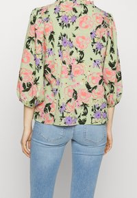 Femme portant un chemisier vert clair avec un motif floral rose et violet et des manches bouffantes trois-quarts, associé à un jean bleu clair.