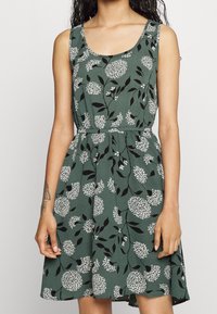 Vihreä hihaton mekko, jossa on valkoinen kukkakuosi, vyötärö on kiristetty ja hame on liehuva. Valmistettu kevyestä kankaasta, jossa on tekstuuripinta.