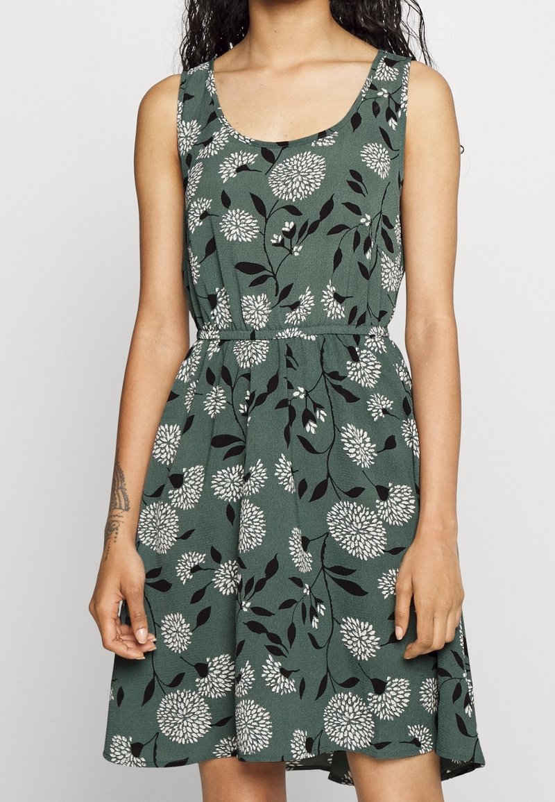 Vihreä hihaton mekko, jossa on valkoinen kukkakuosi, vyötärö on kiristetty ja hame on liehuva. Valmistettu kevyestä kankaasta, jossa on tekstuuripinta.