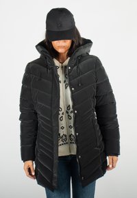 Doudoune noire avec capuche, présentant un design matelassé en chevrons, une fermeture éclair et des cordons de serrage réglables. Portée par-dessus un pull à motifs.