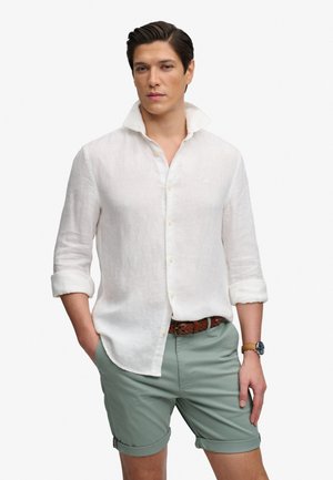 Mężczyzna w białej lnianej koszuli z podwiniętymi rękawami oraz szałwiowozielonych szortach z brązowym plecionym paskiem i zegarkiem na rękę, stojący z jedną ręką w kieszeni.