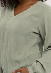 Hellgrünes Langarmoberteil mit V-Ausschnitt, gerippter Textur und einer Fronttasche. Verfügt über geraffte Bündchen an den Ärmeln für zusätzliche Details.