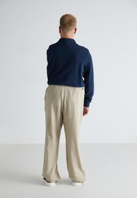 Cămașă cu mânecă lungă navy, pantaloni bej cu croi lejer, textură netedă, talie elastică și adidași albi. Văzut din spate.