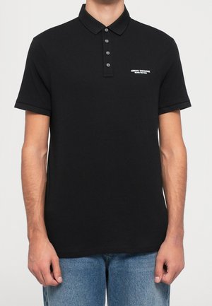 Mężczyzna noszący czarną koszulkę polo Armani Exchange z krótkim rękawem, z trzema guzikami i białym logo na lewej piersi, w połączeniu z niebieskimi dżinsami.