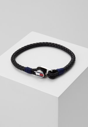 Černý zapletený kožený náramek s akcenty námořnicky modrých nití a stříbrnou sponou s kulatým logem v červené, bílé a modré barvě.
