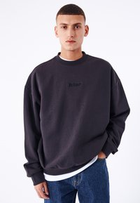 Sudadera negra de gran tamaño con cuello redondo y costuras visibles. Presenta un logotipo bordado en el pecho y mangas caídas.