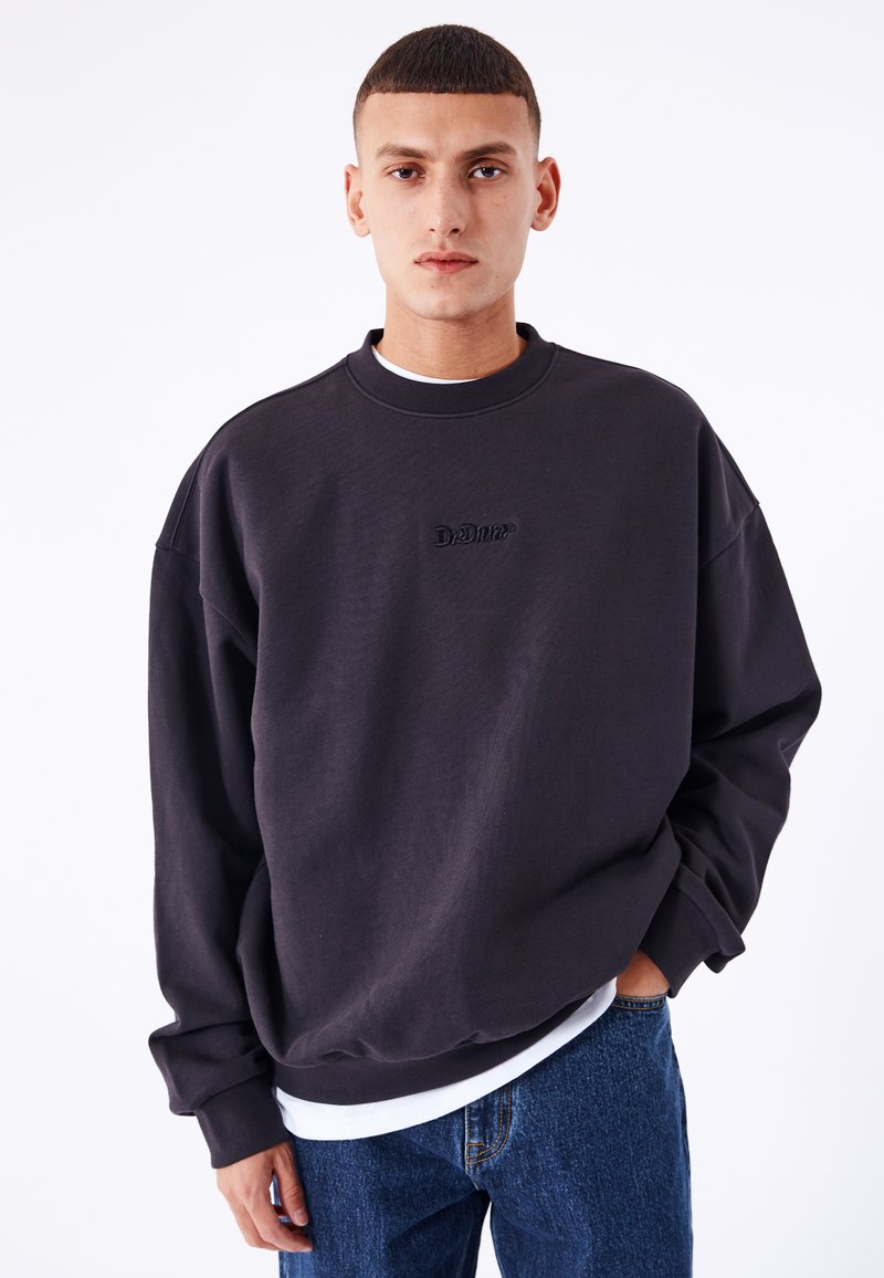 Sudadera negra de gran tamaño con cuello redondo y costuras visibles. Presenta un logotipo bordado en el pecho y mangas caídas.