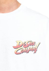 Bílý bavlněný tričko s multicolorovaným grafickým logem "DC SHOE COMPANY" v tučném, dynamickém písmu na levém hrudníku.