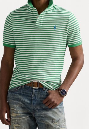 Groen en wit gestreepte polo met kraag, met korte mouwen en een blauw logo. G gedragen met versleten blauwe jeans en een leren bruine riem.