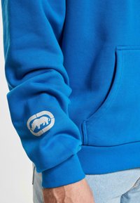 Hoodie bleu en tissu texturé. Présente une poche avant et un logo sur la manche. Les poignets et l'ourlet côtelés assurent un ajustement confortable.
