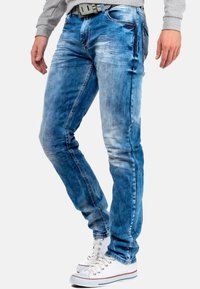 Jean en denim bleu avec un effet délavé, coupe slim, coutures visibles et style cinq poches, associés à des baskets blanches.