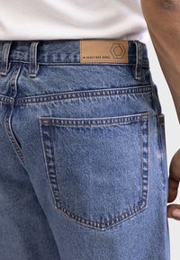 Denim jeans in een middelblauwe tint met een gladde textuur, voorzien van een leren label met de tekst "REDEFINED REBEL" en opvallende stiksels.