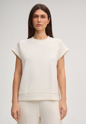 Kobieta o prostych brązowych włosach, ubrana w biały top z krótkim rękawem i białe spodnie, stojąca na tle jasnoszarego tła.