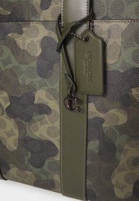 Sac Coach à motif camouflage vert et marron avec une sangle en cuir olive et une étiquette en cuir embossé.
