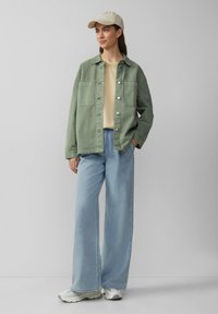 Groene denimjas met zilveren knopen, beige shirt, lichtblauwe wijde jeans en witte sneakers. Aangevuld met een beige pet.