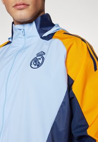 Svijetloplava jakna s patentnim zatvaračem, s tamnoplavim i narančastim detaljima, izrađena od teksturirane tkanine, a na gornjem dijelu prsa nalazi se logo Real Madrida.